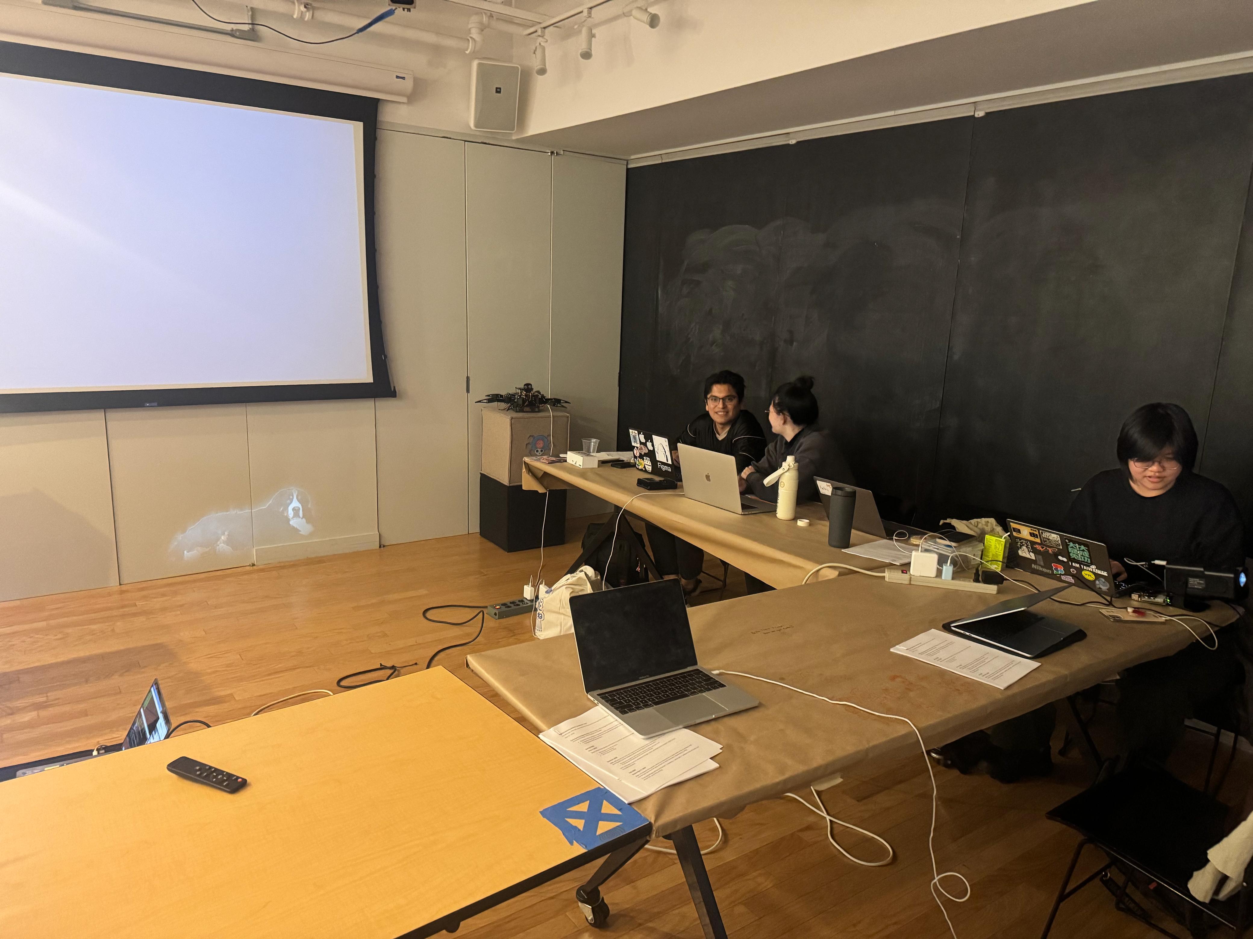 Classroom on break: a projection screen on the left wall begins to light up with Gus the dog while three students sit at the long center table.
